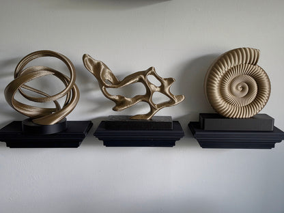 Three decorative wall sculptures displayed on black pedestals against a plain light gray wall: an abstract interlocking ring design, a fluid organic branch-like form, and a spiral ammonite-style shell sculpture.