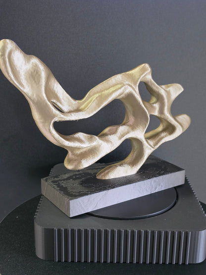Abstract sculptural art piece in a metallic gold finish, black art display base against a dark gray background.