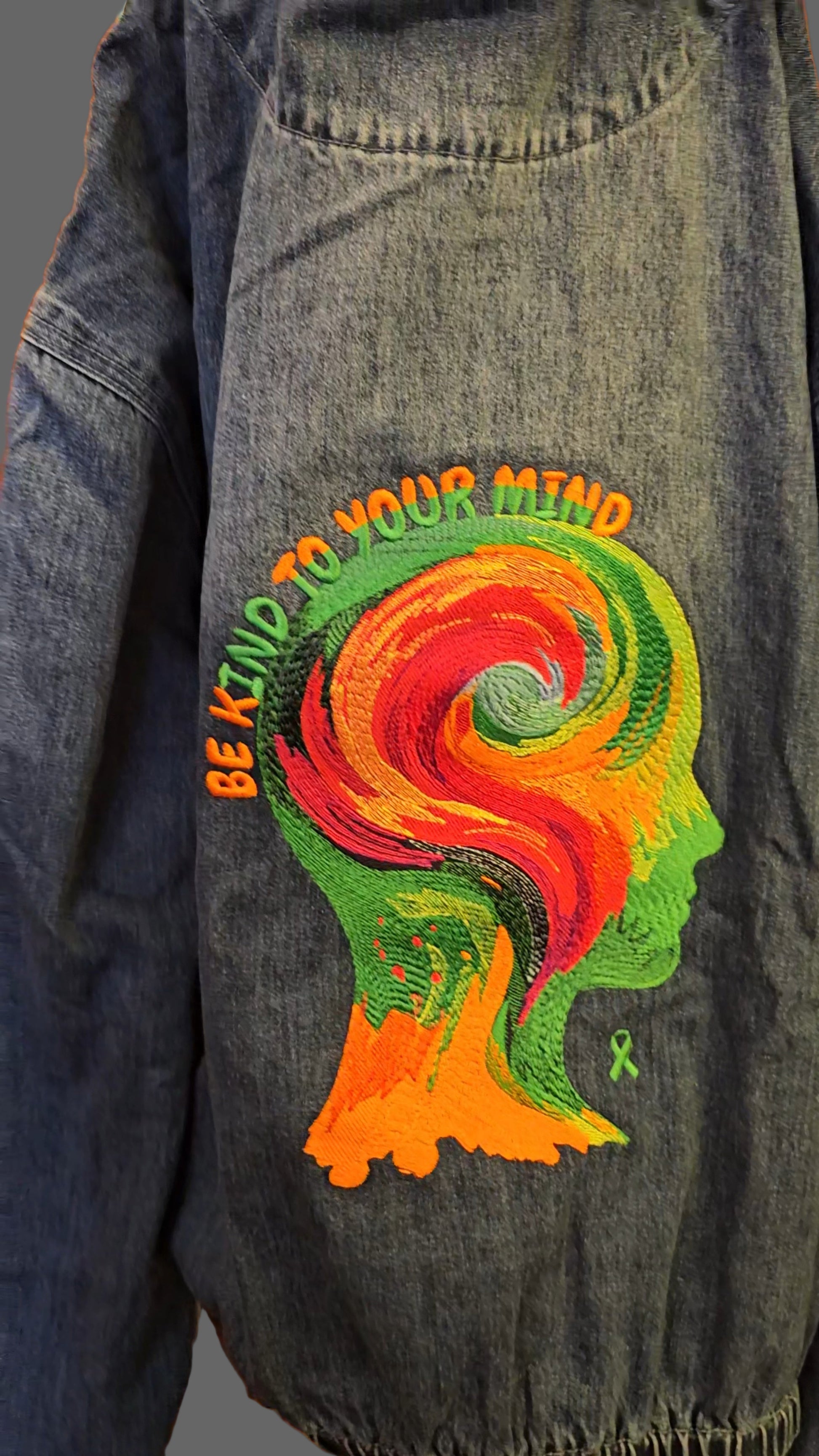 Denim jacket with a colorful embroidered design of a stylized head and 'Be Kind To Your Mind' text.